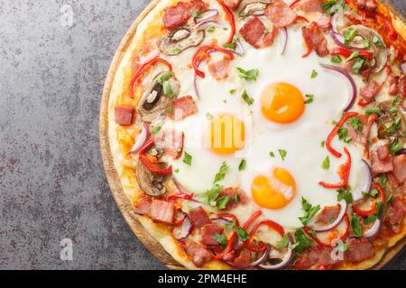 Homemade pizza with bacon, eggs, peppers, mushrooms and red onions close-up on a wooden board on the table. Horizontal top view from above Stock Photo