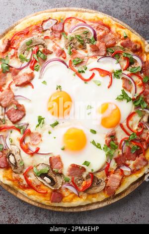Freshly baked pizza with bacon, eggs, peppers, mushrooms, mozzarella and red onions close-up on a wooden board on the table. Vertical top view from ab Stock Photo