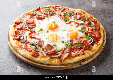 Hot pizza with bacon, eggs, peppers, mushrooms, mozzarella and red onions close-up on a wooden board on the table. Horizontal Stock Photo