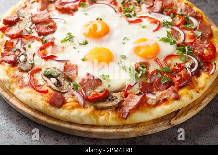 Homemade pizza with bacon, eggs, peppers, mushrooms and red onions close-up on a wooden board on the table. Horizontal Stock Photo