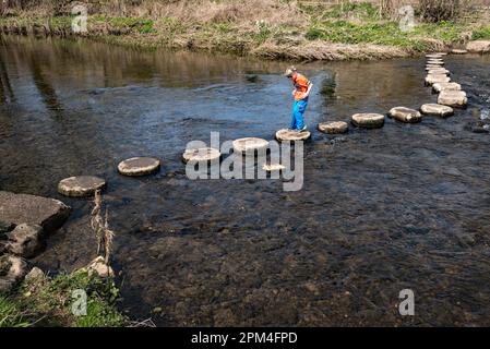 Young boy enjoying crossing the stepping stones in the village of Gargrave in North Yorkshire (Craven area). Stock Photo