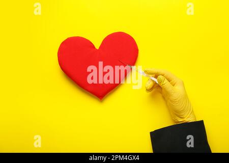 Woman in rubber glove with syringe and heart pillow on yellow background Stock Photo