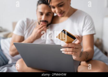 selective focus of credit card and laptop near blurred interracial couple thinking during online shopping in bedroom at home,stock image Stock Photo