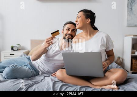 happy bearded man holding credit card near laughing african american girlfriend sitting with laptop on bed at home,stock image Stock Photo