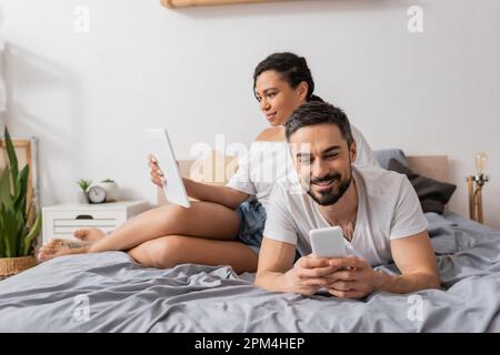 carefree interracial couple in white t-shirts using gadgets while resting on bed at home,stock image Stock Photo