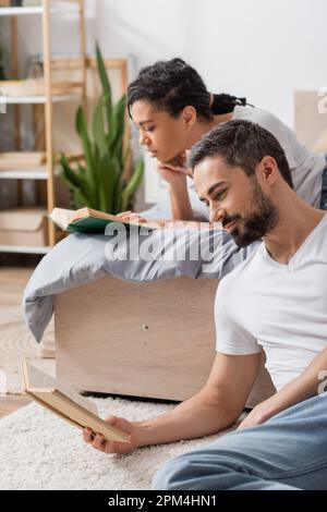 pleased bearded man reading book near blurred african american woman in bedroom at home,stock image Stock Photo