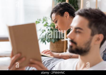 concentrated african american woman reading book near blurred boyfriend in bedroom at home,stock image Stock Photo