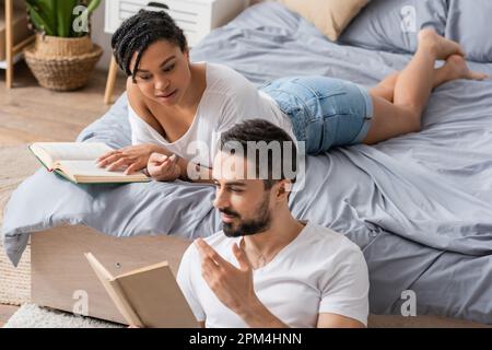 bearded man gesturing and reading book to african american girlfriend lying on bed at home,stock image Stock Photo