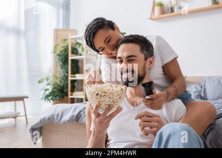 happy african american woman with tv remote controller taking popcorn near smiling bearded man in bedroom at home,stock image Stock Photo