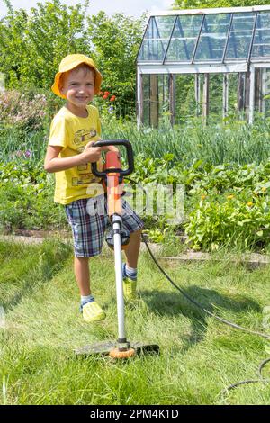 Portrait a boy with an electric lawn mower mowing the lawn. Beauty boy pruning and landscaping a garden, mowing grass, lawn, paths. A boy learns to mo Stock Photo