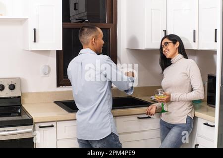 A young man doing housework in the kitchen while talking with his wife. Stock Photo