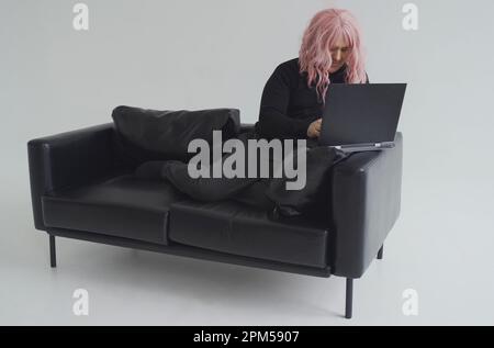 Portrait of a man in a pink wig, sitting on a sofa, typing on a laptop. White background. Stock Photo
