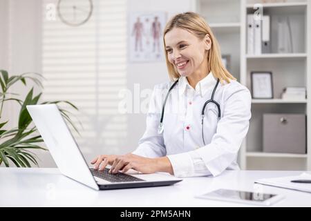 Mature female doctor working inside medical office, pediatrician using laptop while sitting inside clinic office, female worker in medical gown smiling. Stock Photo