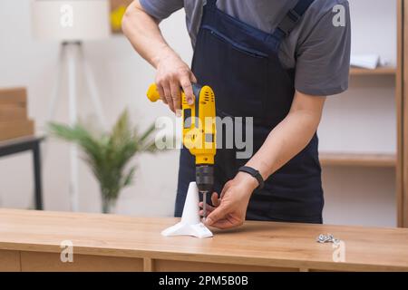 skilled carpenter in blue overalls, using yellow electrical drill screwdriver,Furniture assembling. Stock Photo