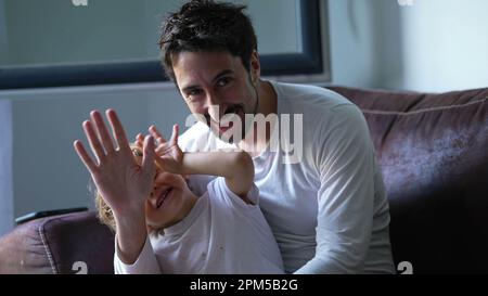 Father and child waving hello to camera sitting on sofa indoors. Dad and little boy saying HI with hand. Parenting lifestyle concept Stock Photo