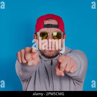 Man with beard,red cap and sun glasses on pointing his fingers in front of himself with a blue background. model release available. Stock Photo