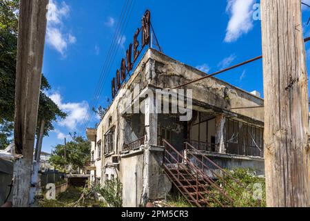 A red, partly weathered advertising sign rises above the ruins of a commercial building in the ghost town of Varosha near Famagusta, Turkish Republic of Northern Cyprus (TRNC). Rusting stair railings and shattered window fronts mark the building. Until the Turkish invasion of 1974, Varosha was a glamorous seaside resort with an international jet set; it has been a military restricted zone ever since, and a small part has been open to visitors since late 2020. Stock Photo