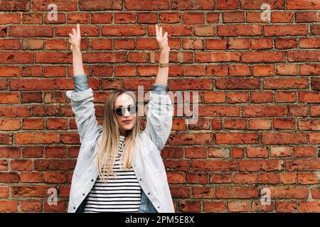 A woman in sunglasses with her hands raised against a brick wall Stock Photo