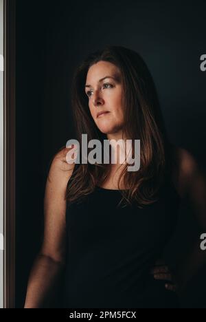 Portrait of attractive woman with long brown hair against dark wall. Stock Photo