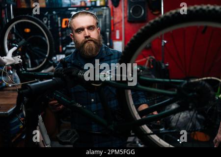 Portrait of handsome bearded cycling repairman with fashion haircut standing by bicycle in repair workshop with dark interior, looking at camera. Stock Photo
