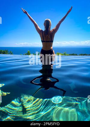 Women in infinity pool with arms outstretched with view of ocean Stock Photo
