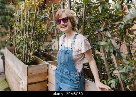 Fashionable woman smiling with sunglasses leaning against plants Stock Photo