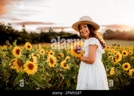 Young  girl with a hat on holding a sunflower in a field at sunset Stock Photo