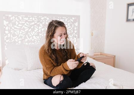 Happy young woman looking at phone sitting on white bed Stock Photo