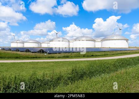 Row of solar panels in front of a group of fuel storage tanks in a business park on a sunny summer day. Wind turbines are visible in background. Stock Photo