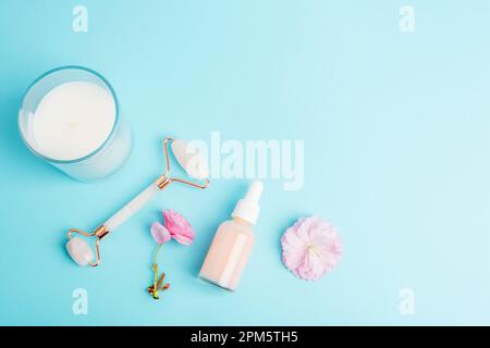 Serum bottle, face roller, gua sha stone, aroma candle and pink sakura blossom on blue background. Skin care, beauty treatment concept. Top view, flat Stock Photo