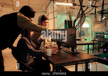 Diverse group of hackers working together to crack server, planting virus to steal personal data at night. Young criminals using monitors to hack computer network and system. Stock Photo