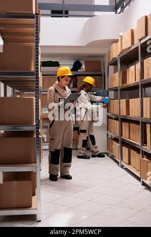 Diverse storehouse workers managing goods stock supply and packing cardboard boxes. Young woman warehouse operative controlling distribution and filling in inventory report Stock Photo