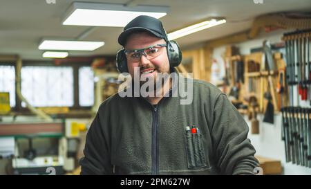 Ready to work male carpenter in protective glasses and headphones smiling at camera. Interior of woodworking workshop. High quality photo Stock Photo