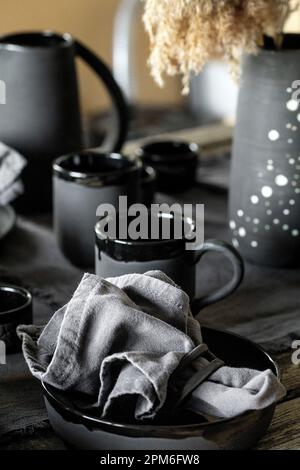 Rustic table setting with empty craft handmade ceramic tableware, black bowls, plates, cups, jug and vase on linen tablecloth. Blck ceramic roses flow Stock Photo