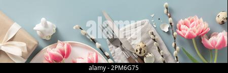 Banner. Table setting. A fashionable minimalistic plate with a linen napkin, Knife and fork, gift box, Easter eggs, feathers and tulips on a blue back Stock Photo