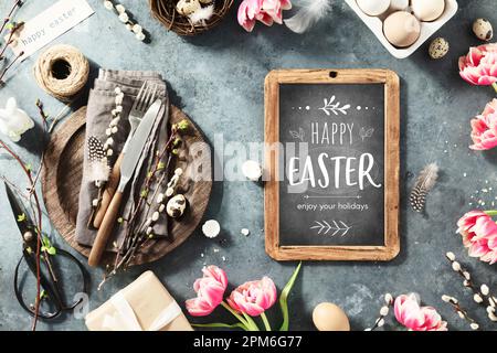 Easter table setting with Happy Easter text on chalk board, spring flowers and cutlery on dark blue background top view flat lay Stock Photo