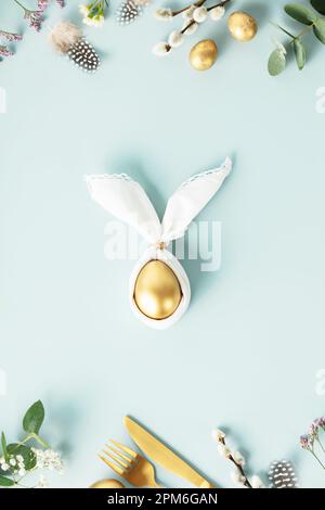 Stylish Easter flat lay with golden egg in easter bunny napkin, golden quail eggs, feathers, cutlery and spring flowers on blue background. Minimalist Stock Photo