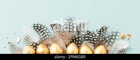 Raw of golden quail eggs and quail feathers on blue background. Beautiful modern minimalistic Easter composition. Copy space, top view, flat lay Stock Photo