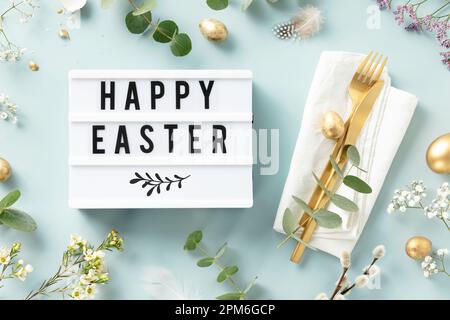 Easter table decorations. Stylish Easter brunch table setting with lightbox text Happy Easter, eggs, vintage cutlery, nests and spring branches on blu Stock Photo