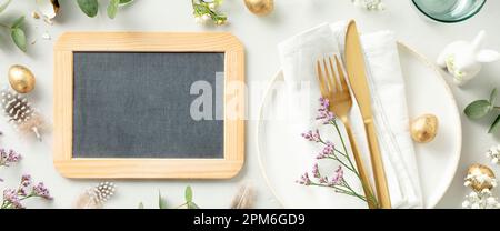 Easter table decorations. Stylish Easter brunch table setting with chalkboard, white plate, napkin, golden cutlery, easter eggs and spring branches on Stock Photo