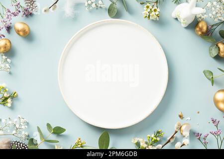 Easter table decorations. Happy Easter concept with white plate, golden easter eggs, feathers and spring flowers on blue background. Flat lay top view Stock Photo
