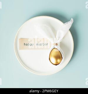 Stylish Easter flat lay with golden egg in easter bunny napkin on white plate over blue background. Minimalist modern Easter table decorations. Top vi Stock Photo