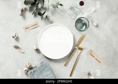 Banner. Table setting. A fashionable minimalistic plate with a linen napkin, Knife and fork, Easter eggs and feathers on a gray background. Top view. Stock Photo