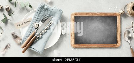 Banner. Table setting. A fashionable minimalistic plate with a linen napkin, knife, fork, chalkboard, Easter eggs and feathers on a gray background. T Stock Photo