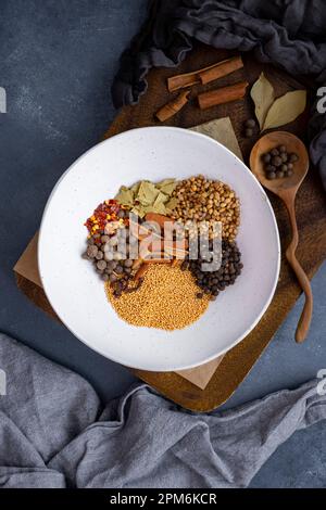 Mustard seeds, coriander seeds, cloves, cinnamon sticks, bay leaves and red pepper flakes mixed in a white ceramic bowl on a wooden board. Stock Photo