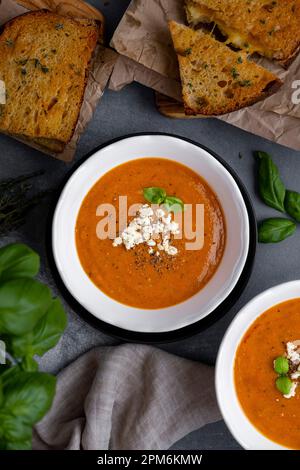 Roasted tomato soup topped with feta cheese and basil leaves in two white bowls and grilled cheese sandwiches on the side. Stock Photo