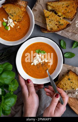 Woman hands holding a bowl of tomato soup and the spoon inside it, another bowl of tomato soup and grilled cheese sandwiches on the side. Stock Photo