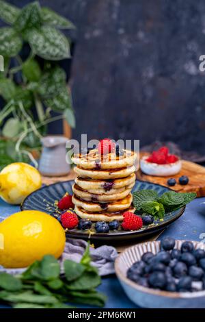 A stack of pancakes garnished with raspberries, blueberries and fresh mint on a dark backdrop. Lemons, blueberries and raspberries around them. Stock Photo