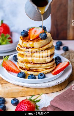 Maple syrup being poured over a stack of pancakes garnished with blueberries and strawberries. Stock Photo