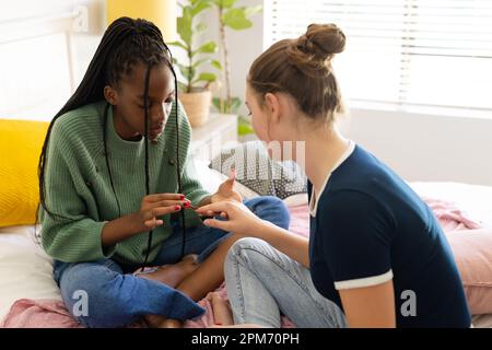 Happy diverse teenager girls friends sitting on bed and painting nails Stock Photo